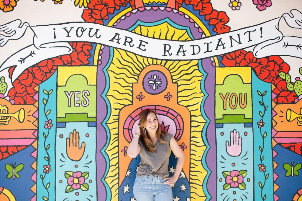 Becky Launder laughing in front of a neon sign reading You Are Radiant